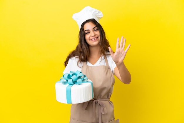 Mulher jovem chef de pastelaria caucasiana com um grande bolo isolado em um fundo amarelo e contando cinco com os dedos