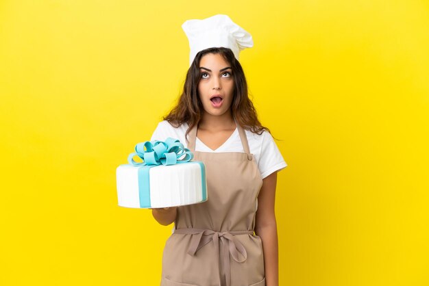 Mulher jovem chef de pastelaria caucasiana com um grande bolo isolado em um fundo amarelo, olhando para cima e com expressão de surpresa