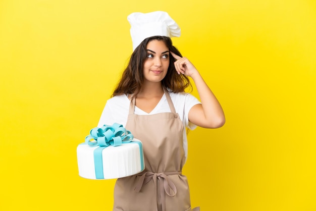 Mulher jovem chef de pastelaria caucasiana com um grande bolo isolado em um fundo amarelo, tendo dúvidas e pensando