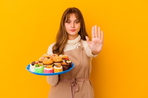 Mulher jovem chef de pastelaria isolada na parede amarela em pé com a mão estendida, mostrando o sinal de pare, impedindo você.