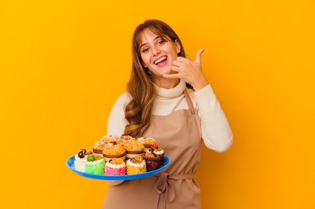 Mulher jovem chef de pastelaria isolada na parede amarela, mostrando um gesto de chamada de telefone móvel com os dedos.