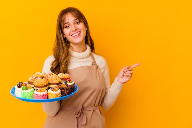 Mulher jovem chef de pastelaria isolada na parede amarela, sorrindo e apontando de lado, mostrando algo no espaço em branco.