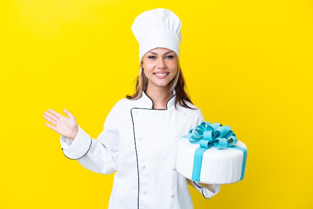 Mulher jovem chef de pastelaria russa com um bolo grande isolado em fundo amarelo, estendendo as mãos para o lado para convidar para vir