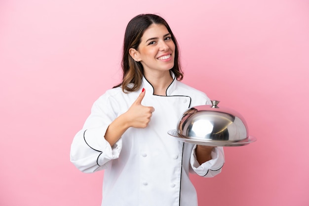 Mulher jovem chef italiano segurando a bandeja com tampa isolada no fundo rosa, dando um polegar para cima gesto