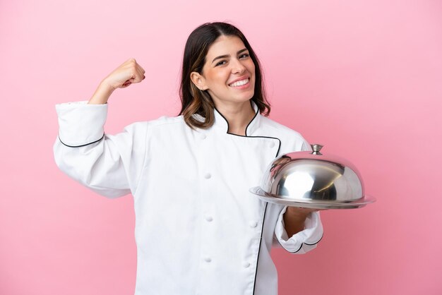 Mulher jovem chef italiano segurando a bandeja com tampa isolada no fundo rosa, fazendo um gesto forte