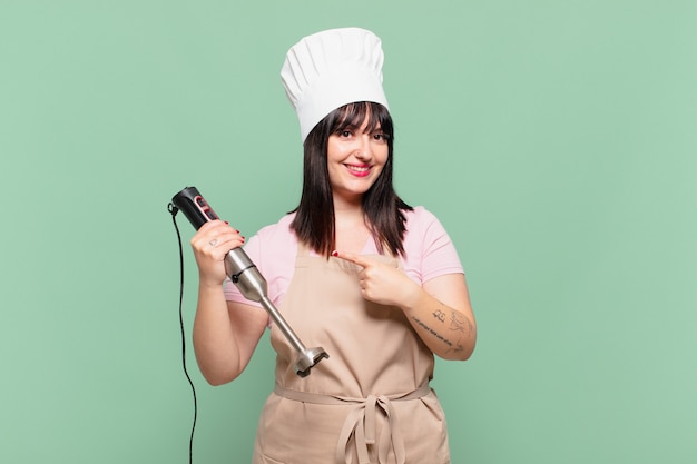 Mulher jovem chef sorrindo alegremente, sentindo-se feliz e apontando para o lado e para cima, mostrando o objeto no espaço da cópia