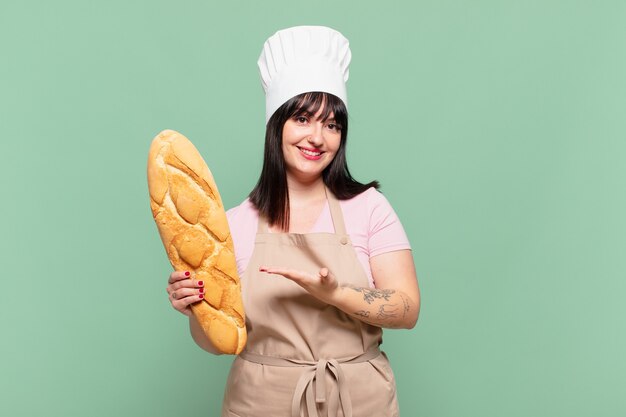 Mulher jovem chef sorrindo alegremente, sentindo-se feliz e mostrando um conceito no espaço da cópia com a palma da mão