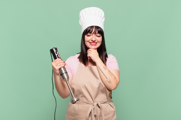 Mulher jovem chef sorrindo com uma expressão feliz e confiante com a mão no queixo, pensando e olhando para o lado