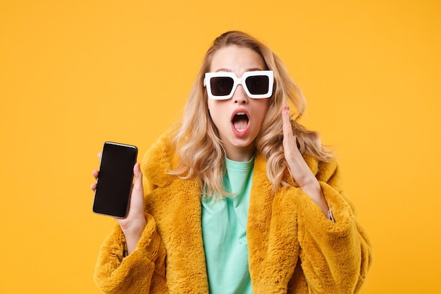 Mulher jovem chocada com casaco de pele amarelo, óculos escuros posando isolado em fundo laranja. Conceito de estilo de vida de pessoas. Mock up espaço de cópia. Segure o celular com a tela vazia em branco, espalhando as mãos.