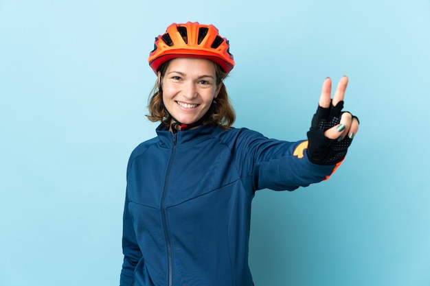 Mulher jovem ciclista isolada na parede azul sorrindo e mostrando o sinal da vitória