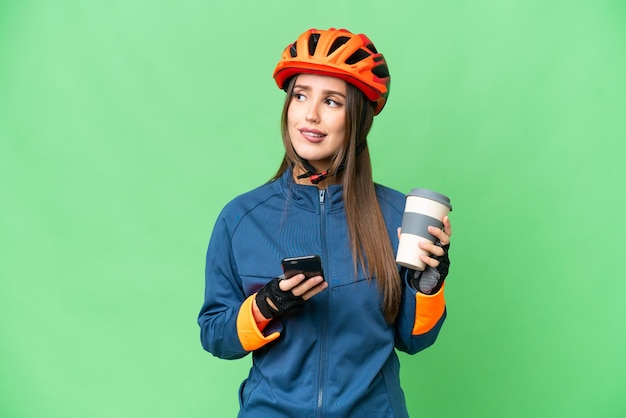 Mulher jovem ciclista sobre fundo croma isolado segurando café para levar e um celular enquanto pensava em algo