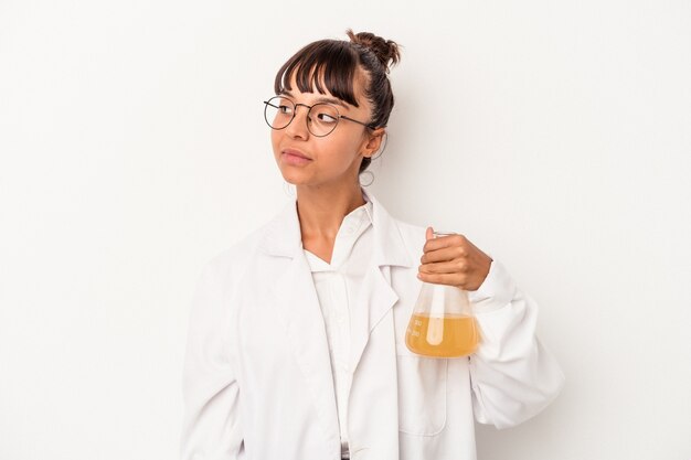 Mulher jovem cientista de raça mista segurando um tubo de ensaio isolado no fundo branco parece de lado sorrindo, alegre e agradável.