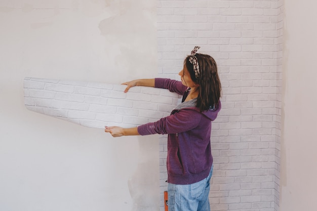 Foto mulher jovem cola papel de parede em casa. garota cola papel de parede na forma de um tijolo. reparar.