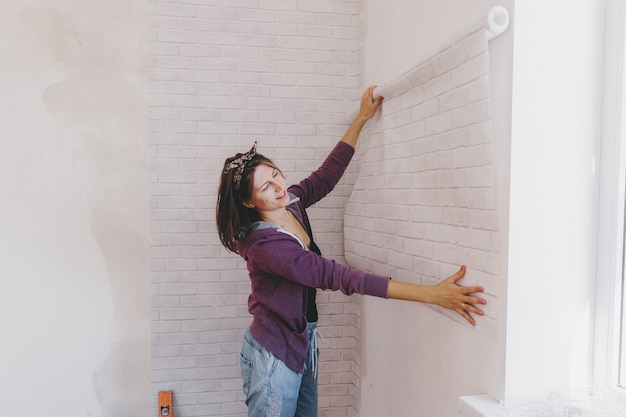 Mulher jovem cola papel de parede em casa. garota cola papel de parede na forma de um tijolo. reparar.