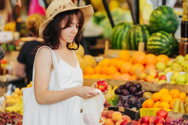 Mulher jovem coloca frutas e vegetais em uma sacola de produtos de algodão no mercado de alimentos. Saco ecológico reutilizável para fazer compras. Conceito de desperdício zero.