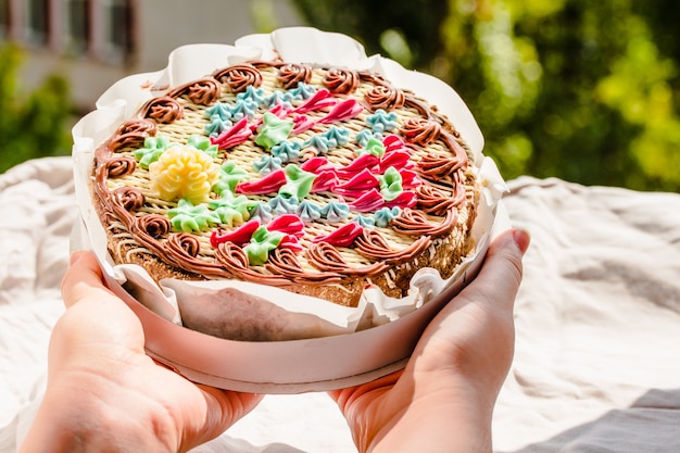 Mulher jovem com as mãos segurando o bolo de nozes redondo inteiro recém-inaugurado tradicional Kiev na embalagem. Os produtos de panificação ucranianos conhecidos. Dia de sol e fundo dos verdes.