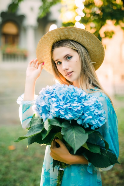 Mulher jovem com buquet de hedragea azul em um vestido azul em pé no jardim verde ao pôr do sol