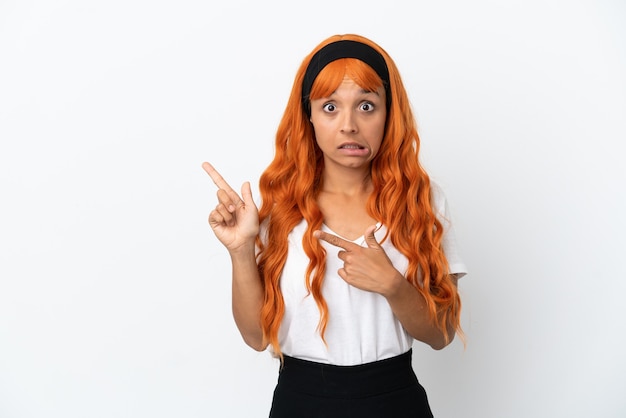 Mulher jovem com cabelo laranja isolado no fundo branco assustada e apontando para o lado