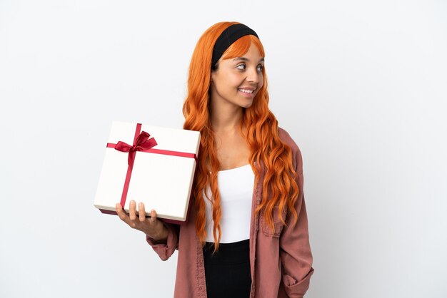 Mulher jovem com cabelo laranja segurando um presente isolado no fundo branco, olhando para o lado e sorrindo