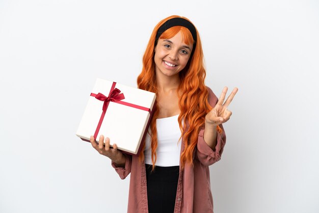Mulher jovem com cabelo laranja segurando um presente isolado no fundo branco, sorrindo e mostrando o sinal da vitória