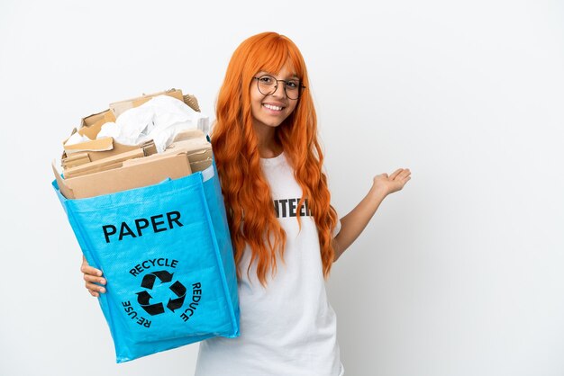 Mulher jovem com cabelo laranja segurando uma sacola de reciclagem cheia de papel para reciclar isolada no fundo branco estendendo as mãos para o lado para convidar para vir