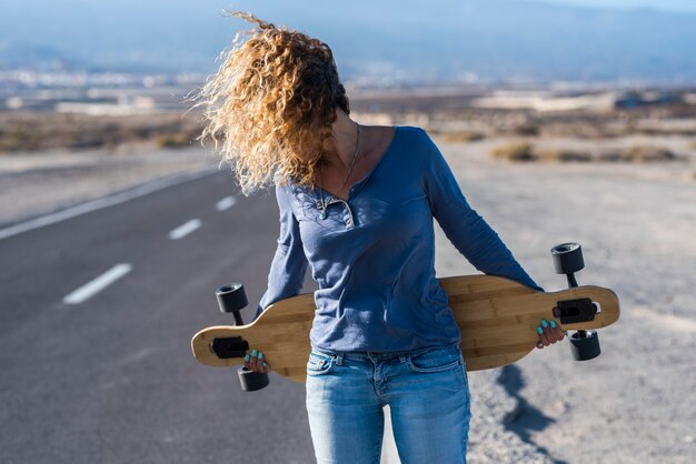 Mulher jovem com cabelo movido pelo rosto escondido do vento e longa prancha de skate andar livre em viagens de lazer ao ar livre com uma longa estrada de asfalto no fundo - mulheres desfrutam de um estilo de vida ativo