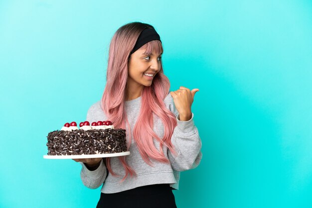 Mulher jovem com cabelo rosa segurando um bolo de aniversário isolado em um fundo azul apontando para o lado para apresentar um produto