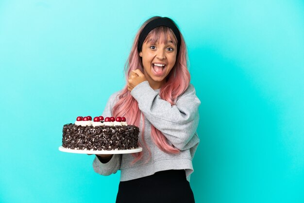 Mulher jovem com cabelo rosa segurando um bolo de aniversário isolado em um fundo azul e comemorando uma vitória