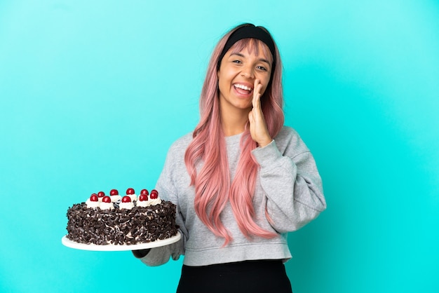 Mulher jovem com cabelo rosa segurando um bolo de aniversário isolado em um fundo azul e gritando com a boca aberta
