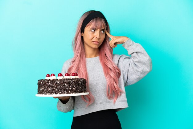 Mulher jovem com cabelo rosa segurando um bolo de aniversário isolado em um fundo azul, tendo dúvidas e pensando
