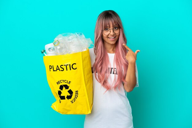 Mulher jovem com cabelo rosa segurando uma sacola cheia de garrafas de plástico para reciclar isolada em um fundo azul fazendo um gesto de polegar para cima