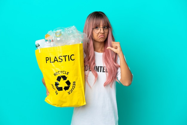 Mulher jovem com cabelo rosa segurando uma sacola cheia de garrafas de plástico para reciclar isolada em um fundo azul orgulhosa e satisfeita