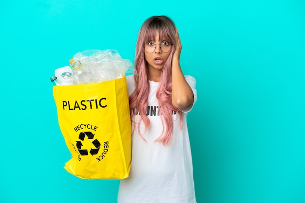 Foto mulher jovem com cabelo rosa segurando uma sacola cheia de garrafas plásticas para reciclar isolada em um fundo azul com expressão de surpresa