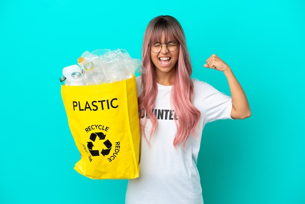 Mulher jovem com cabelo rosa segurando uma sacola cheia de garrafas plásticas para reciclar isolada em um fundo azul fazendo um gesto forte