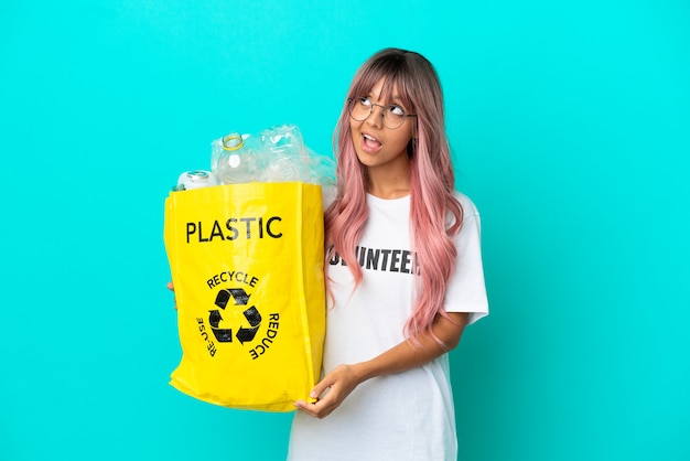 Mulher jovem com cabelo rosa segurando uma sacola cheia de garrafas plásticas para reciclar isolada em um fundo azul tendo uma ideia enquanto olha para cima
