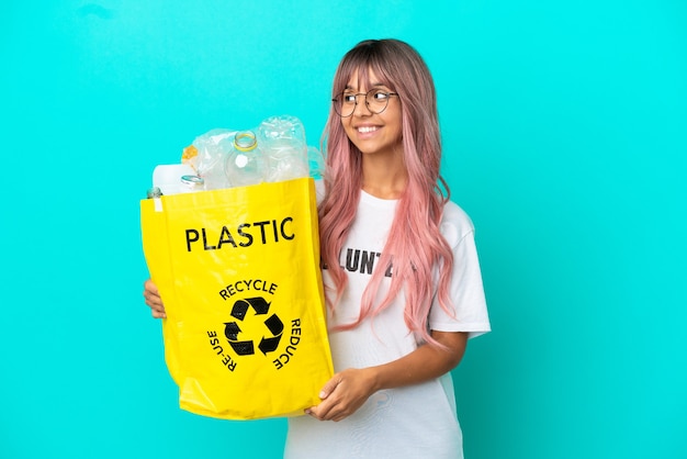 Mulher jovem com cabelo rosa segurando uma sacola cheia de garrafas plásticas para reciclar isoladas em um fundo azul, olhando para o lado