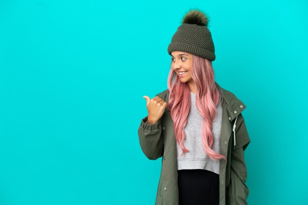 Mulher jovem com cabelo rosa vestindo um casaco à prova de chuva isolado em um fundo azul apontando para o lado para apresentar um produto