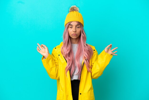 Mulher jovem com cabelo rosa vestindo um casaco à prova de chuva isolado em um fundo azul em pose zen