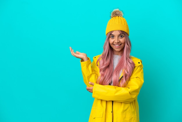 Mulher jovem com cabelo rosa, vestindo um casaco à prova de chuva isolado em um fundo azul, estendendo as mãos para o lado para convidar para vir