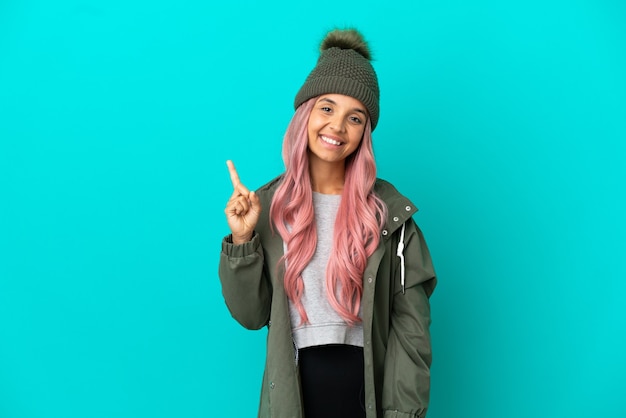 Mulher jovem com cabelo rosa vestindo um casaco à prova de chuva isolado em um fundo azul, mostrando e levantando um dedo em sinal dos melhores