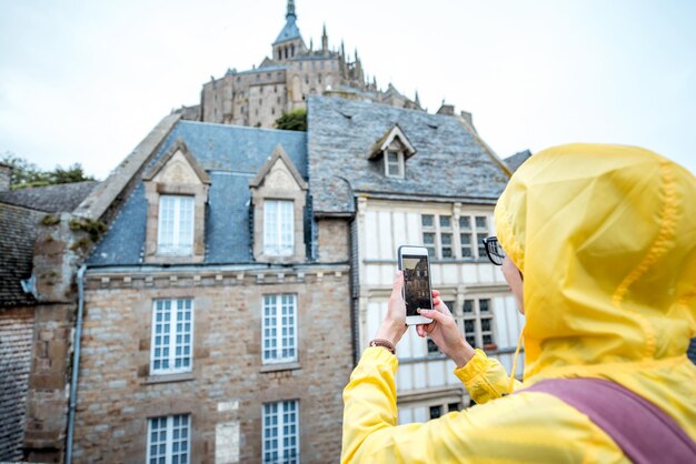 Mulher jovem com capa de chuva amarela fotografando edifícios visitando a famosa ilha do Mont Saint Michel, na França