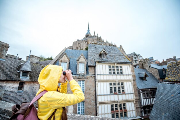 Mulher jovem com capa de chuva amarela observando com binóculos belos edifícios na famosa ilha Mont Saint Michel, na França