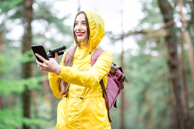 Mulher jovem com capa de chuva amarela usando smartphone para orientação durante a caminhada na floresta de pinheiros verdes