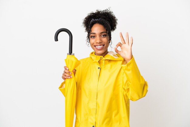 Mulher jovem com casaco à prova de chuva e guarda-chuva isolado no fundo branco, mostrando sinal de ok com os dedos