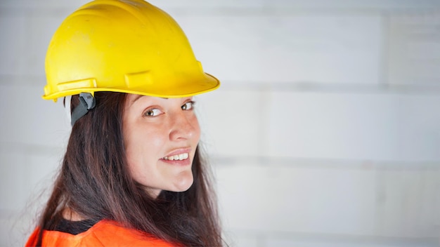 Foto mulher jovem com chapéu amarelo e colete laranja de alta visibilidade cabelo longo e escuro olhando por cima do ombro sorrindo confiante espaço de fundo de parede desfocado do local de construção para o texto do lado direito