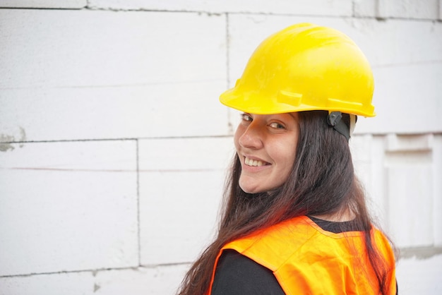 Mulher jovem com chapéu amarelo e colete laranja de alta visibilidade, cabelos longos e escuros, mãos cruzadas, olhando por cima do ombro, sorrindo confiante, fundo borrado na parede do local de construção.