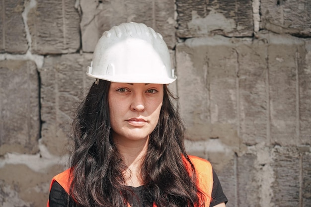 Foto mulher jovem com chapéu branco e colete laranja de alta visibilidade cabelo longo e escuro olhando para a câmera fundo borrado da parede do antigo canteiro de obras