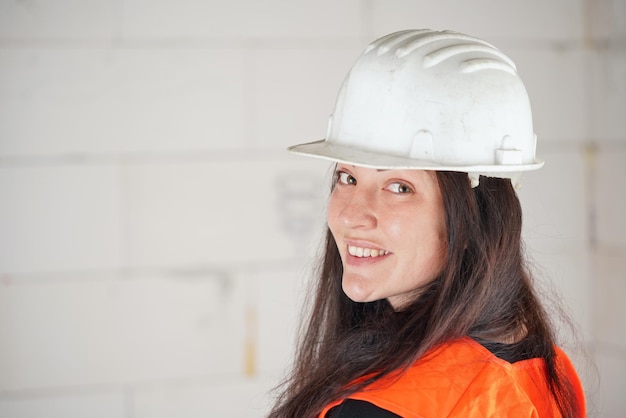 Foto mulher jovem com chapéu branco e colete laranja de alta visibilidade cabelo longo e escuro olhando por cima do ombro para a câmera sorrindo fundo borrado da parede do local de construção