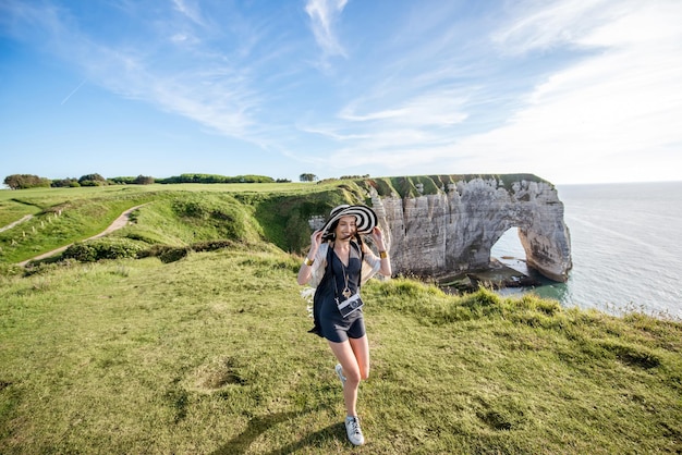 Mulher jovem com chapéu listrado e suéter, desfrutando de uma bela vista da famosa costa rochosa perto da cidade de etretat, na frança