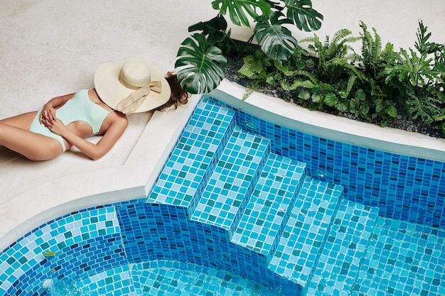 Mulher jovem com corpo em forma perfeita tomando banho de sol perto da piscina com chapéu de palha no rosto, vista de cima